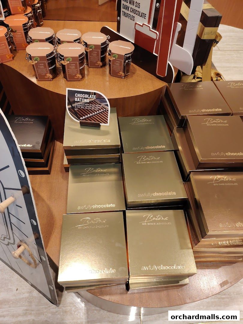 A display of Awfully Chocolate Batons in gold packaging, with jars of chocolate truffles in the background.
