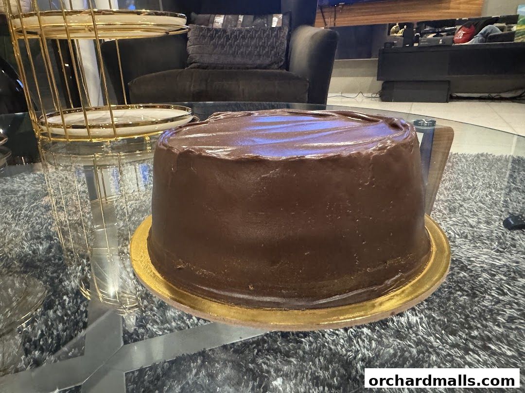 A rich chocolate cake sits on a glass table next to a gold tiered stand.