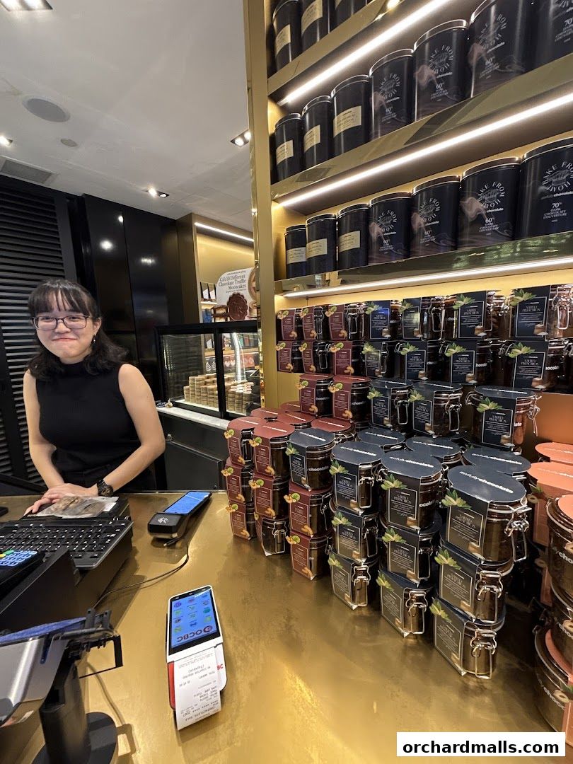 A smiling employee stands behind a counter with shelves of chocolate products and jars.