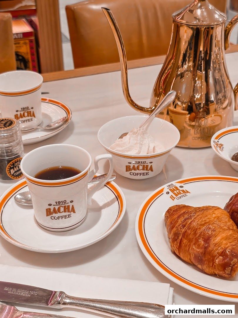 A table setting with Bacha Coffee cups, a golden teapot, and a croissant.