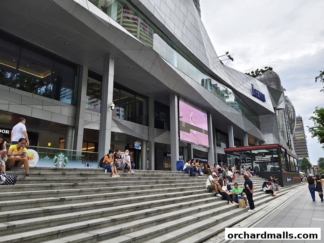 People relaxing on the steps outside a modern shopping mall with a large digital display.