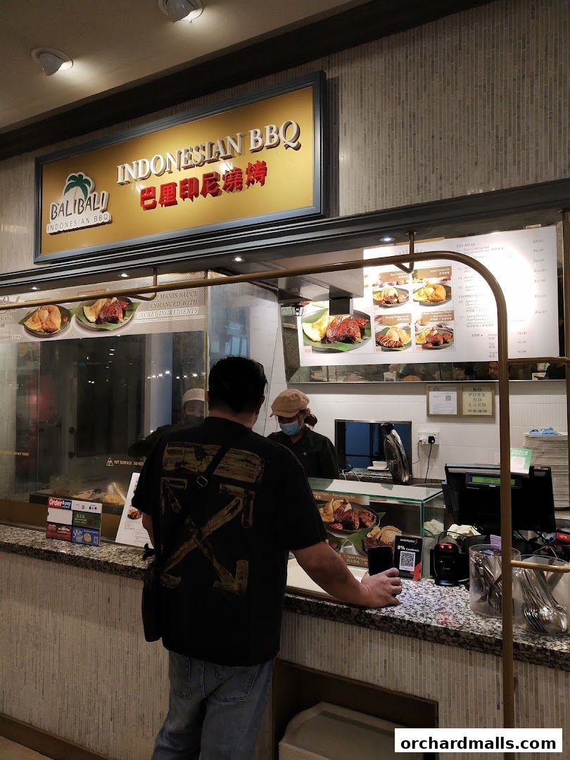 A customer stands at the counter of BaliBali Indonesian BBQ, with the menu and food displays visible.