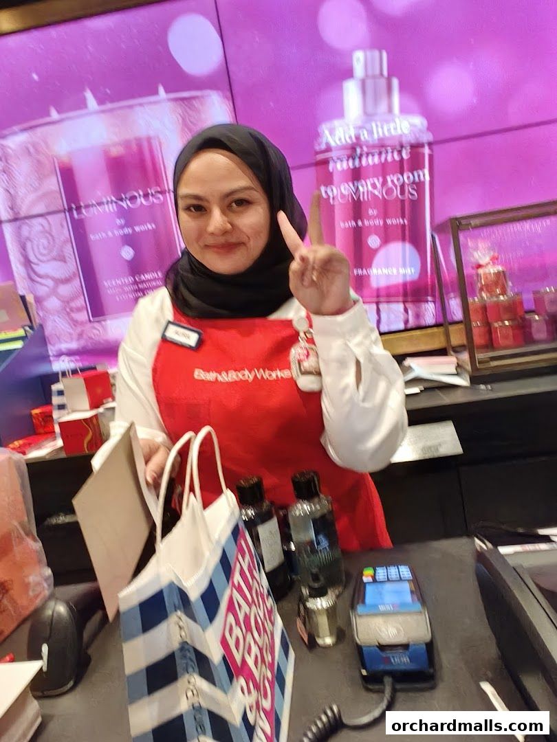 A smiling employee at Bath & Body Works holds up a shopping bag and flashes a peace sign.