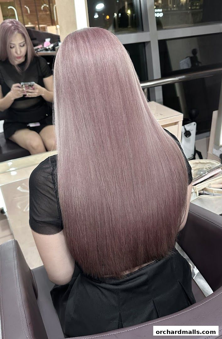 A woman with long, straight, rose-tinted hair sits in a salon chair, reflected in a mirror.