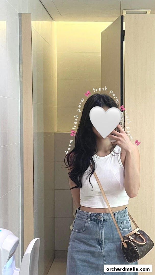 A person with long, wavy dark hair poses in a white tank top and denim skirt.