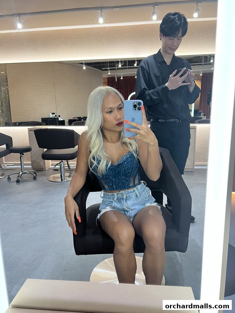 A woman with blonde hair takes a selfie in a salon with a stylist in the background.