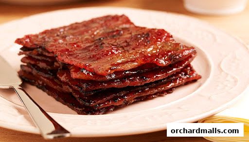 A stack of glistening, sweet bak kwa slices on a pink plate with a serving utensil.