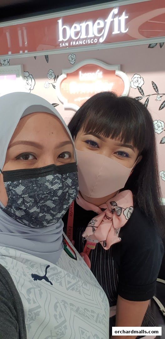 Two women wearing masks pose in front of a Benefit Cosmetics Brow Bar.