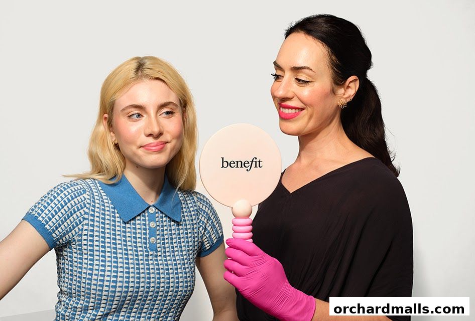 Two women, one with blonde hair and one with dark hair, are looking at a mirror with the Benefit logo.