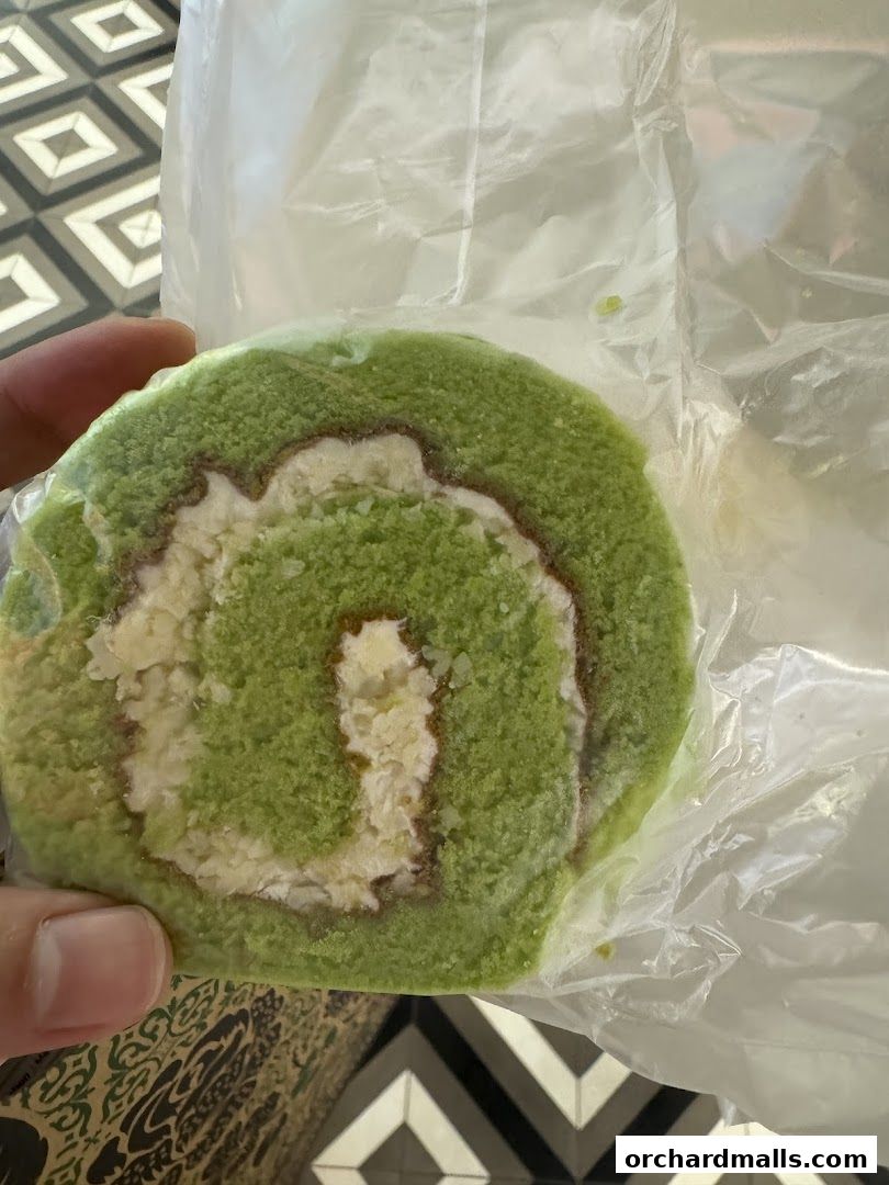 A hand holds a slice of green swiss roll cake with cream filling, wrapped in plastic.