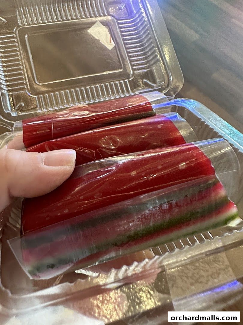 A hand holds a clear plastic container with layered colorful desserts wrapped in plastic.