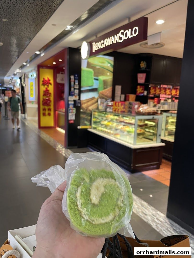 A hand holds a green swiss roll in front of the Bengawan Solo shop.
