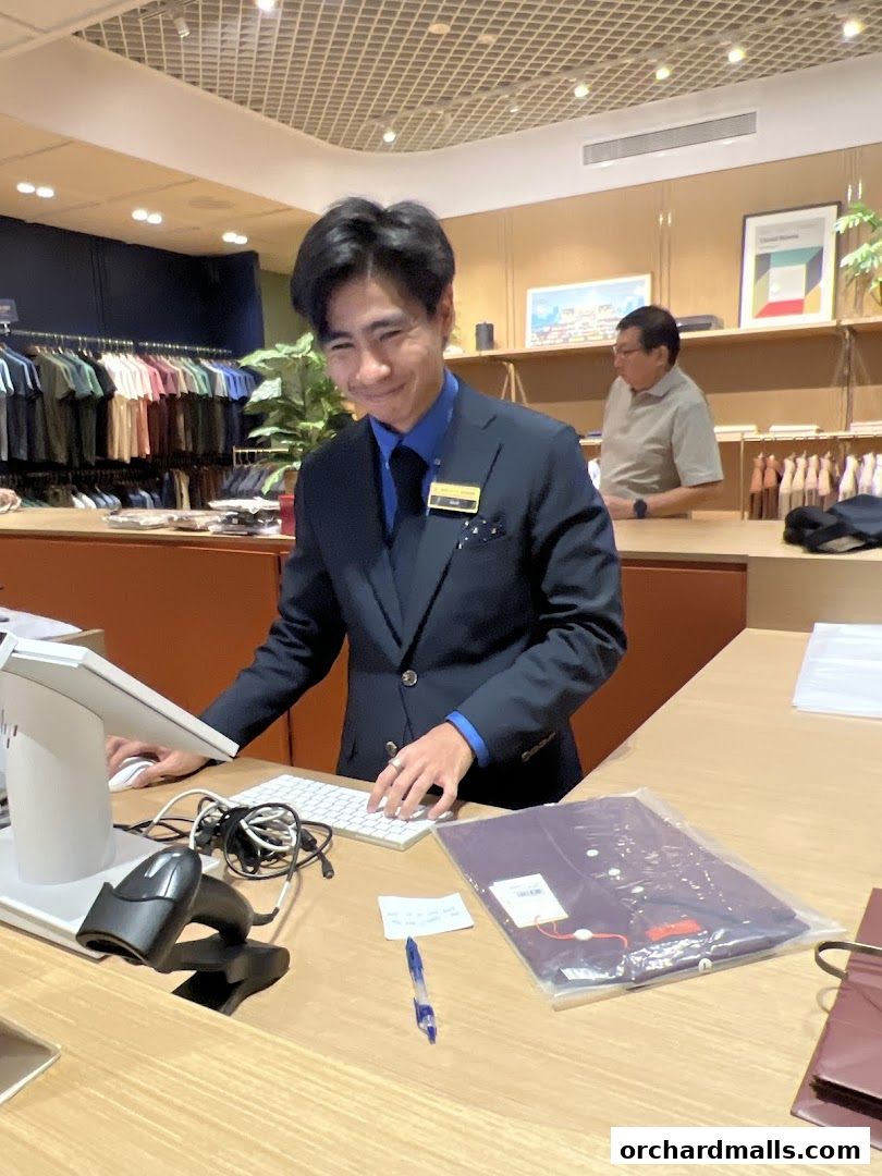 A smiling shop assistant in a suit stands at a counter in a clothing store.