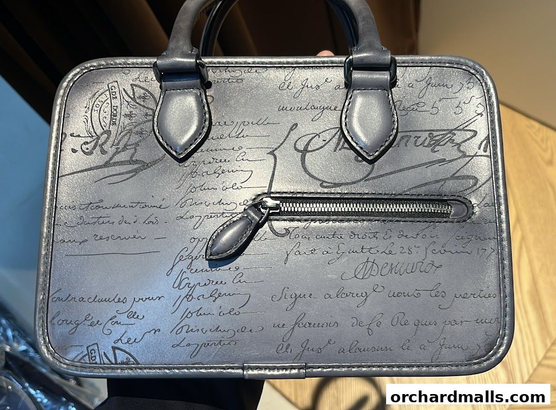 A close-up of a grey Berluti handbag with a script pattern and a zippered pocket.
