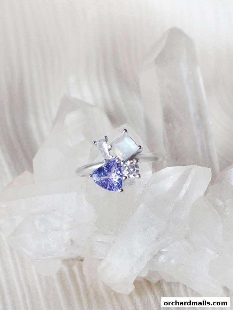 A delicate silver ring with a heart-shaped blue stone and other gems rests on crystals.