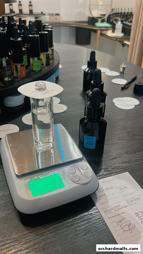 A digital scale measures a vial of liquid next to perfume bottles and a scent recipe card.