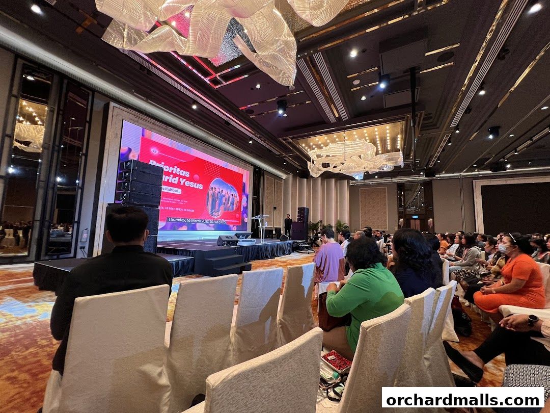 An audience watches a presentation on a large screen in a well-lit conference hall.