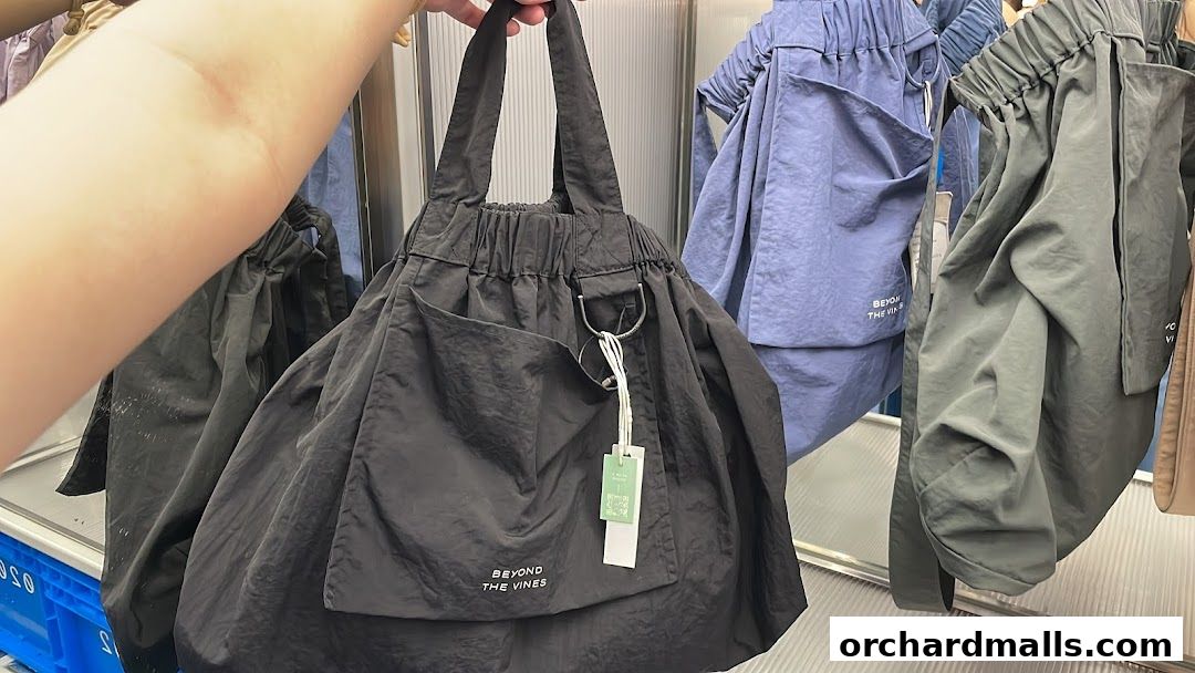 A person holds a black tote bag with other bags in various colors displayed behind.