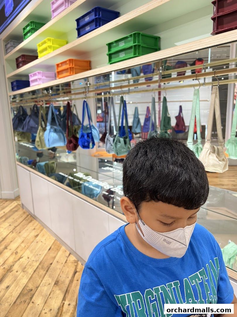 A young boy wearing a mask stands in front of shelves displaying colorful bags and storage crates.