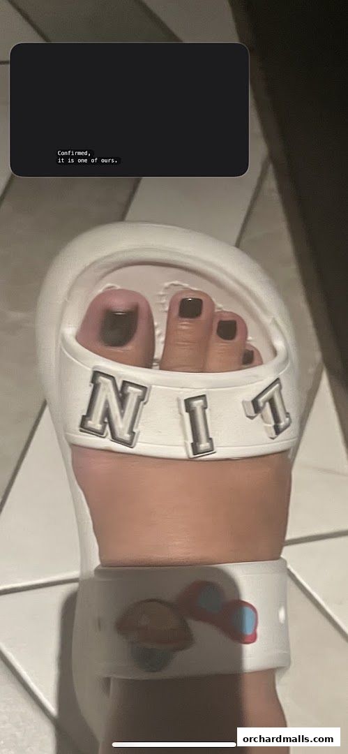A close-up of a foot with dark nail polish wearing white slides with letters.
