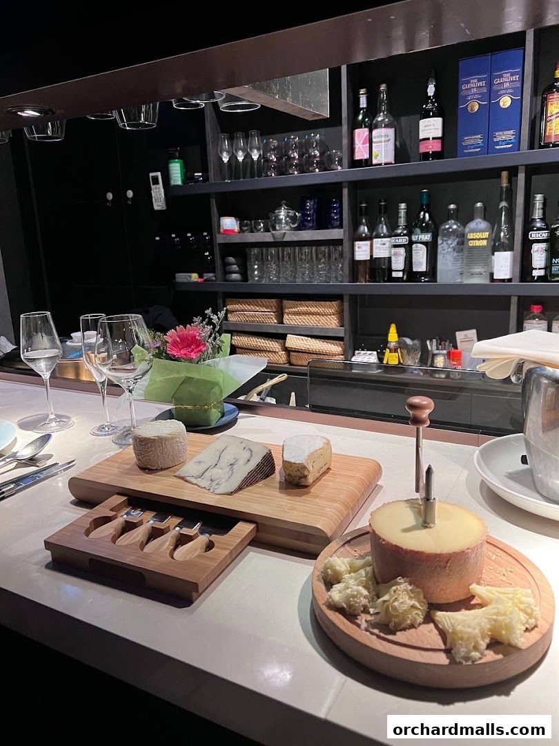 A cheese board with various cheeses and cheese knives, set against a bar with bottles and glassware.