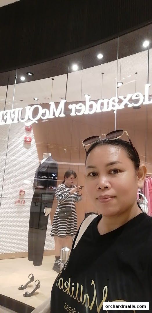 A woman poses in front of a store window displaying a mannequin and clothing.