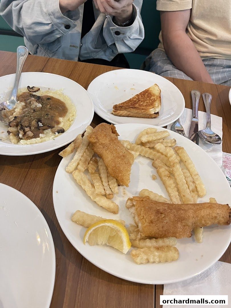 A table with plates of food including fish and chips, grits with mushrooms, and a grilled cheese sandwich.