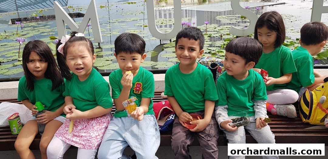 Children in green shirts sitting on a bench with snacks near a water feature.