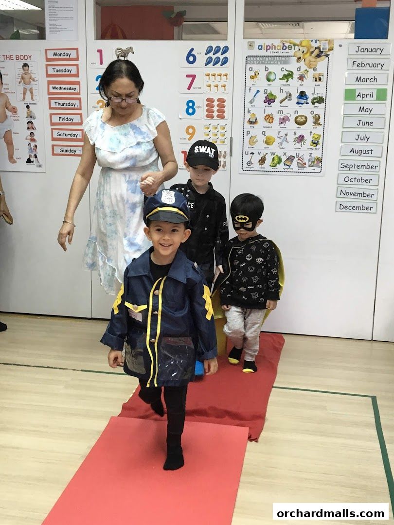 Children in costumes walk on a red carpet with a teacher.