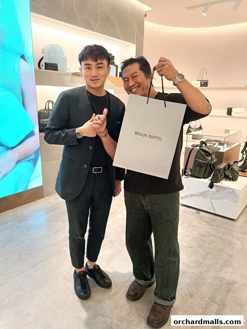 Two men smiling inside a Braun Büffel store, one holding a shopping bag.