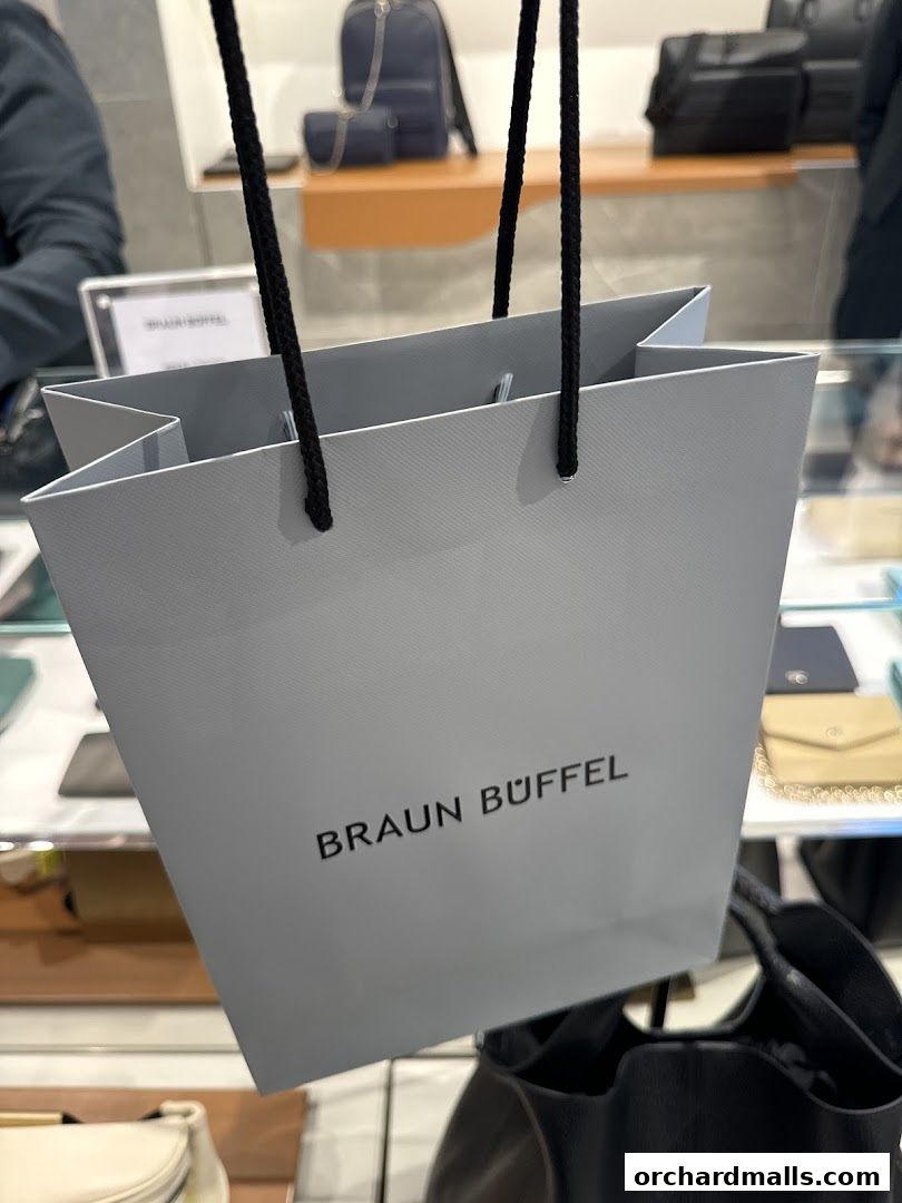 A shopping bag with the Braun Büffel logo is held up in front of a display of bags.