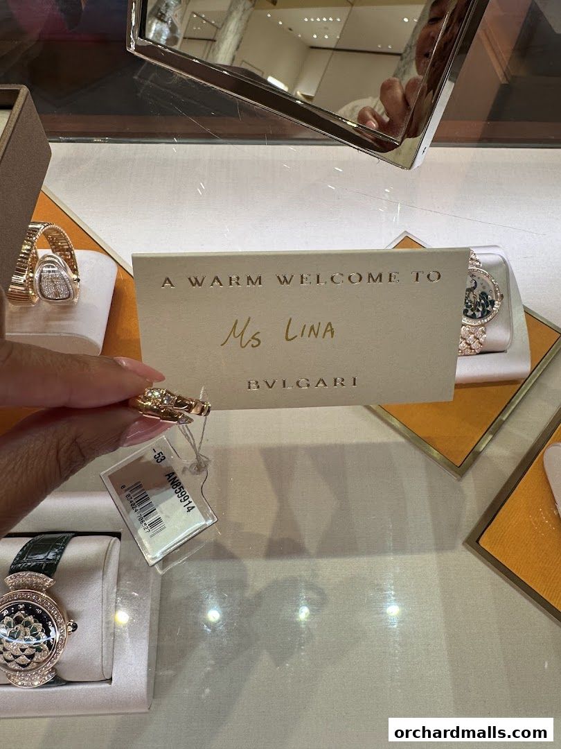A personalized welcome card from Bvlgari is held in front of luxury watches and jewelry in a display case.