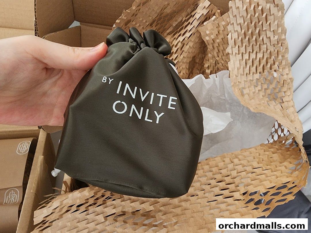 A hand holds a dark green drawstring pouch with 'BY INVITE ONLY' printed in white.