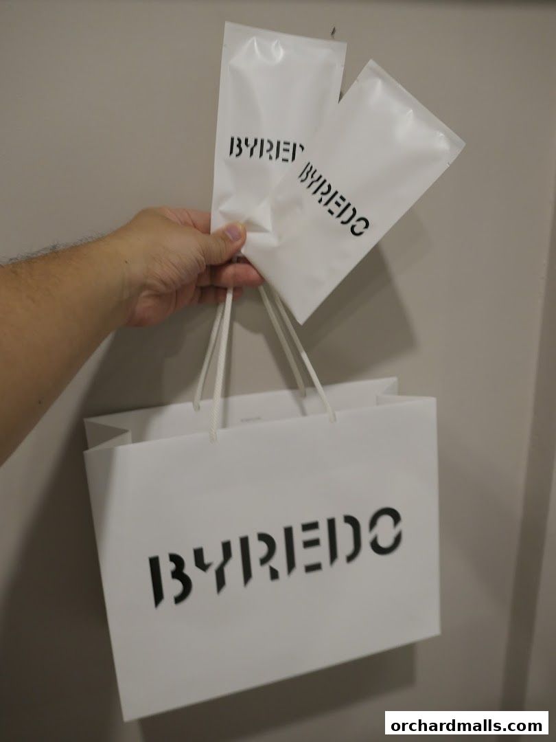 A hand holds a Byredo shopping bag and two smaller Byredo sample packets.