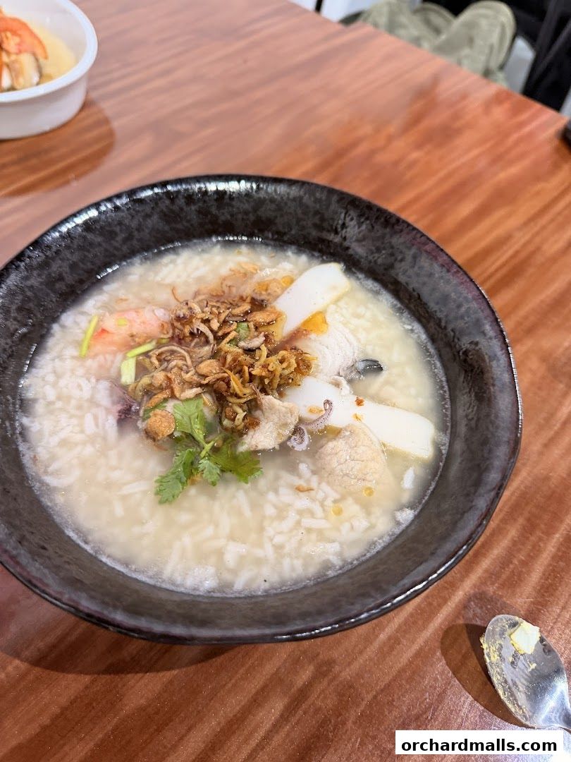 A close-up of a bowl of congee with seafood and toppings on a wooden table.