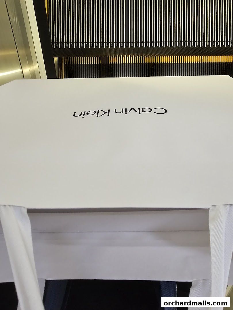 A white shopping bag with the Calvin Klein logo is held up in front of an escalator.