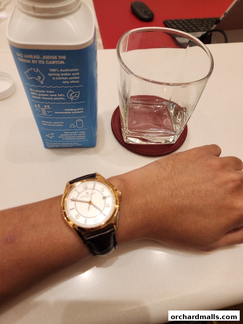 A person's wrist wearing a gold watch, next to a glass of water and a carton of water.