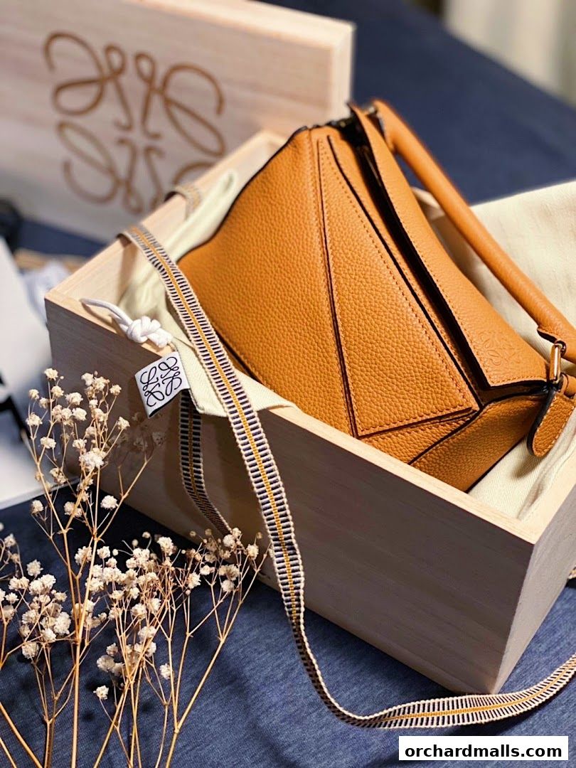 A tan leather Loewe handbag is displayed in a wooden box with dried flowers.