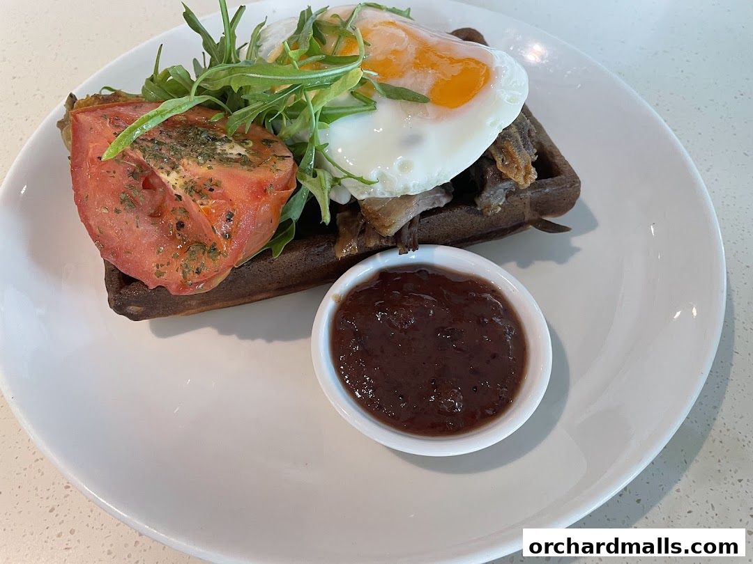 A savory waffle topped with pulled meat, fried egg, tomato, and greens, served with a side of jam.