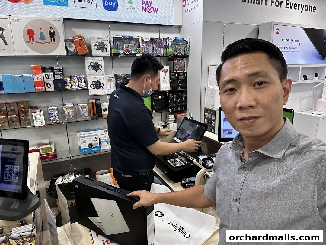 A man in a grey shirt takes a selfie in a tech store with shelves of electronics behind him.