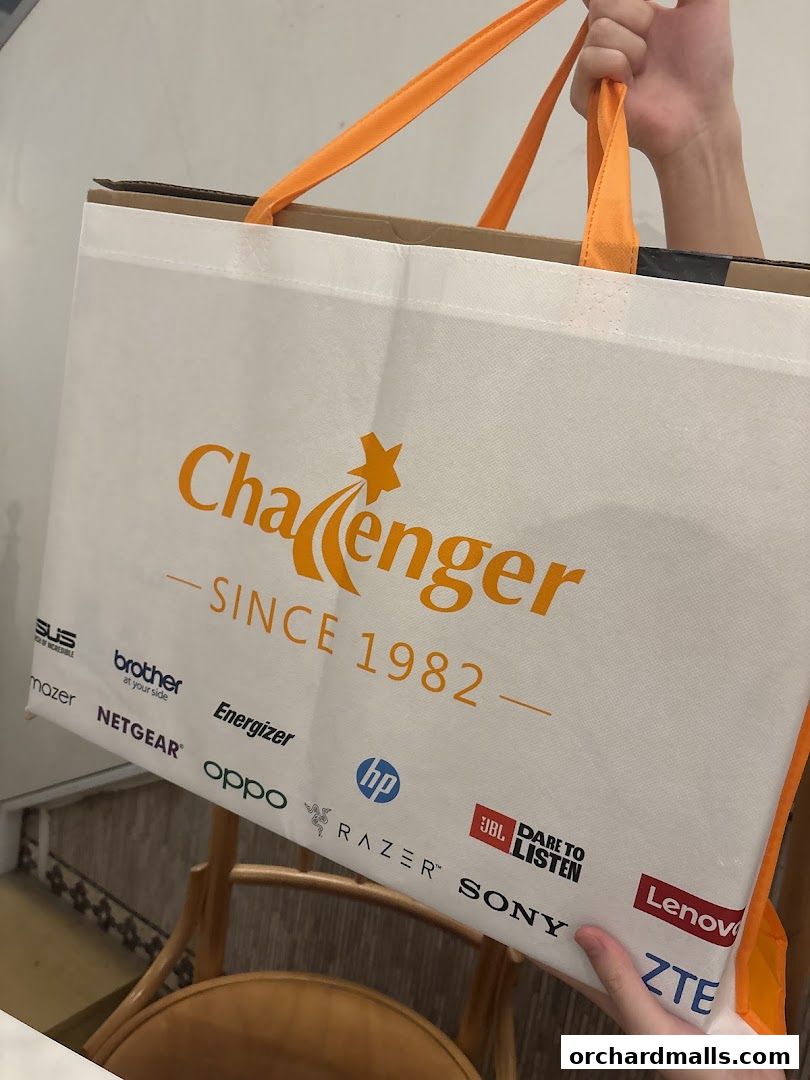 A person holds a white Challenger shopping bag with orange handles, featuring various tech brand logos.
