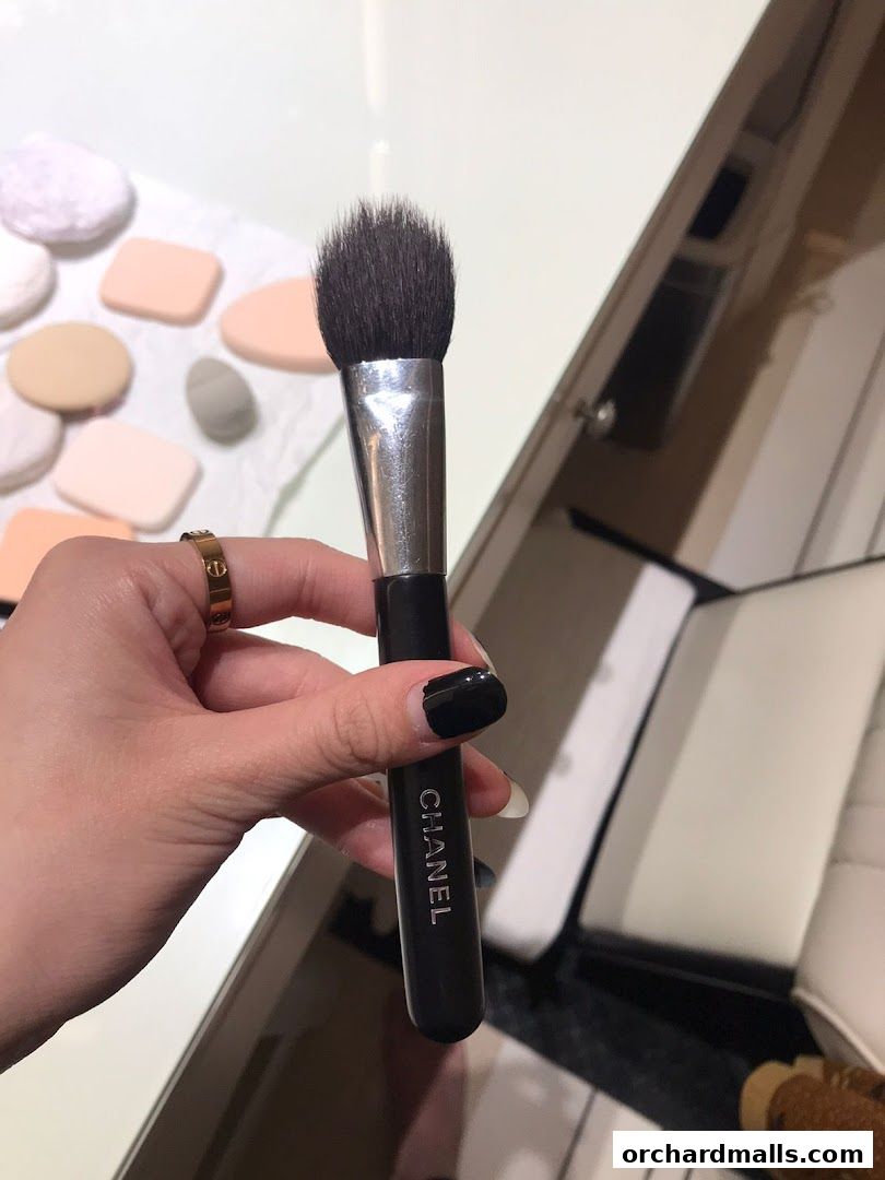 A hand holds a Chanel makeup brush with makeup sponges in the background.