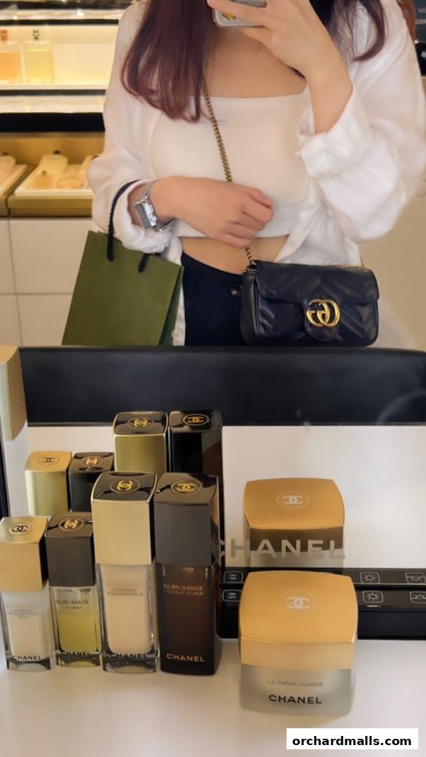 A person holding a Chanel bag and shopping bag in front of Chanel beauty products.