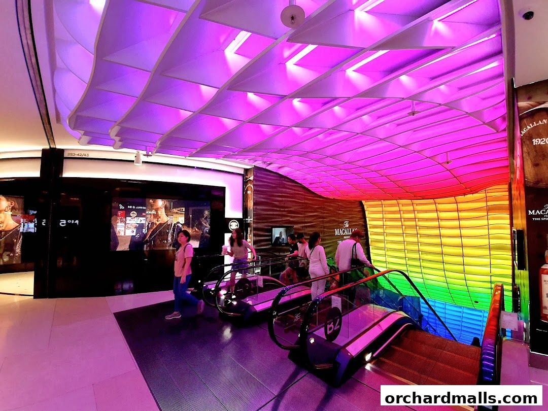 People on escalators under a colorful, illuminated ceiling in a modern shopping mall.