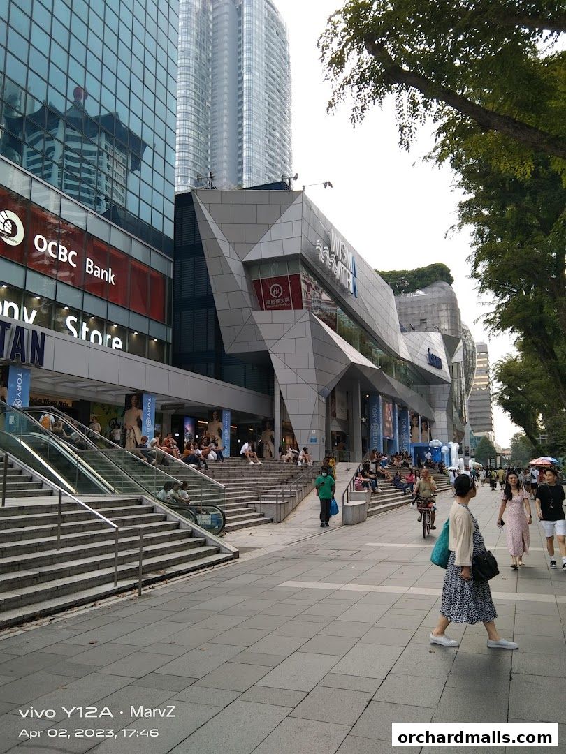 A busy street scene in front of a modern shopping mall with escalators and people walking.