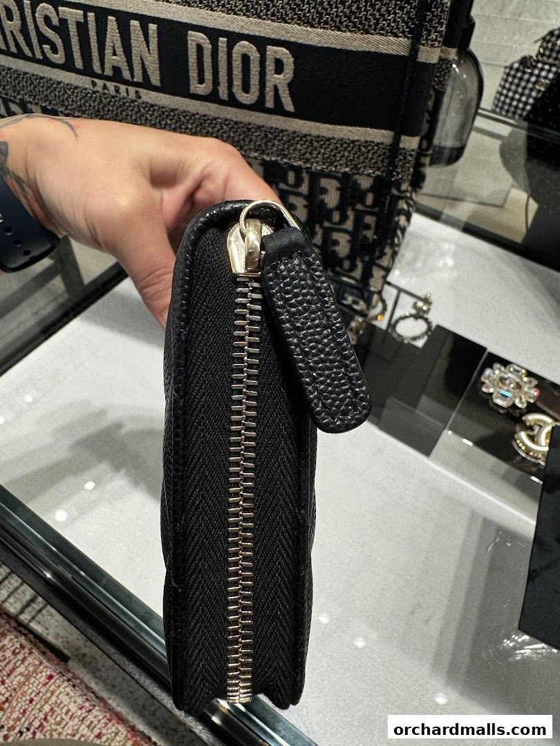 A hand holds a black Chanel wallet with a gold zipper in front of a Dior bag.
