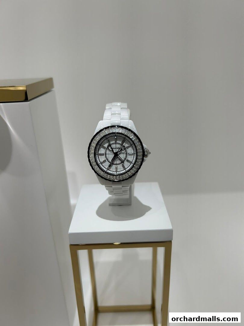 A white Chanel watch with a diamond bezel is displayed on a white pedestal.
