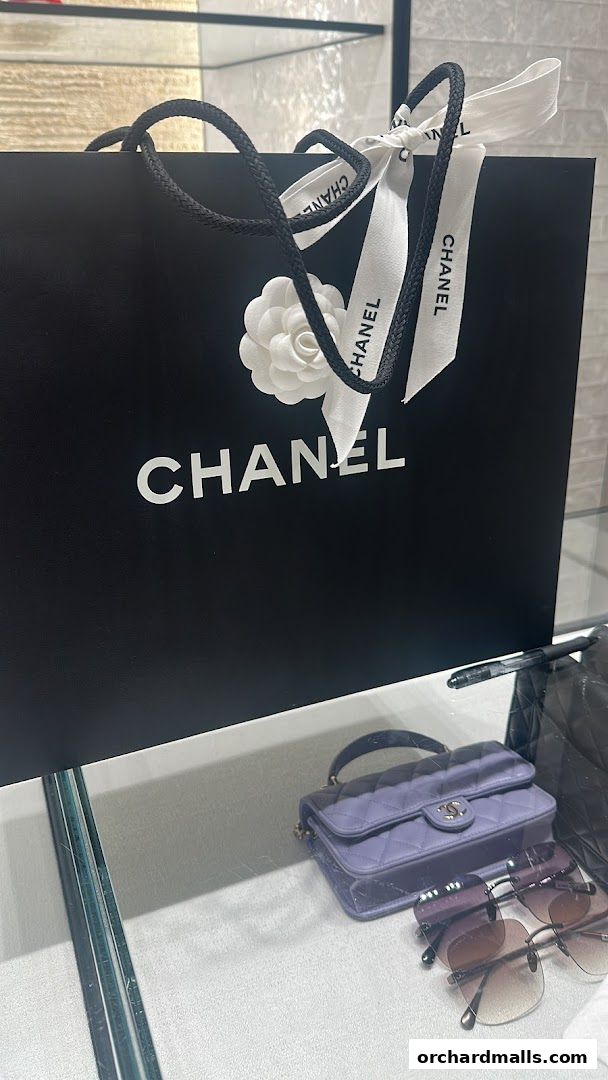 A Chanel shopping bag with a camellia flower and ribbon, next to a purple handbag and sunglasses.