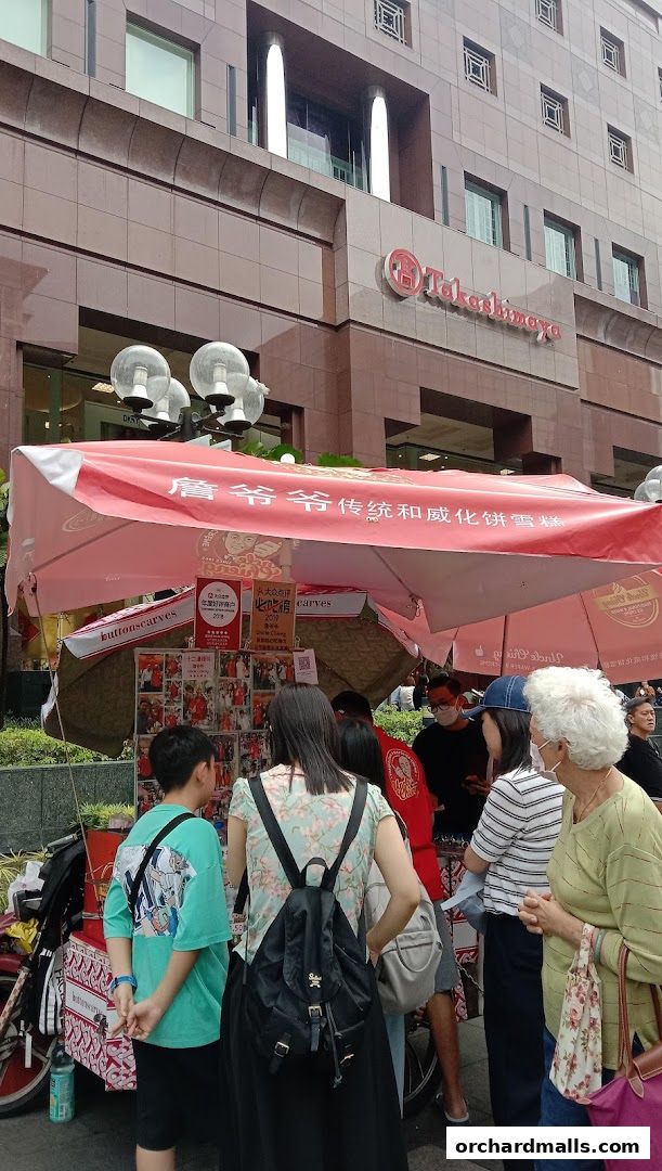 People gathered around a food stall with a red canopy in front of a department store.