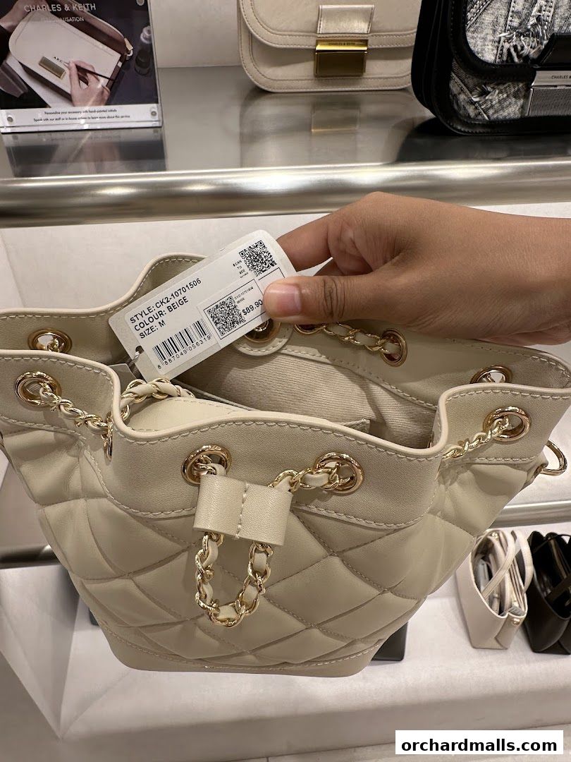 A hand holds a tag on a quilted beige bucket bag with gold chain details.